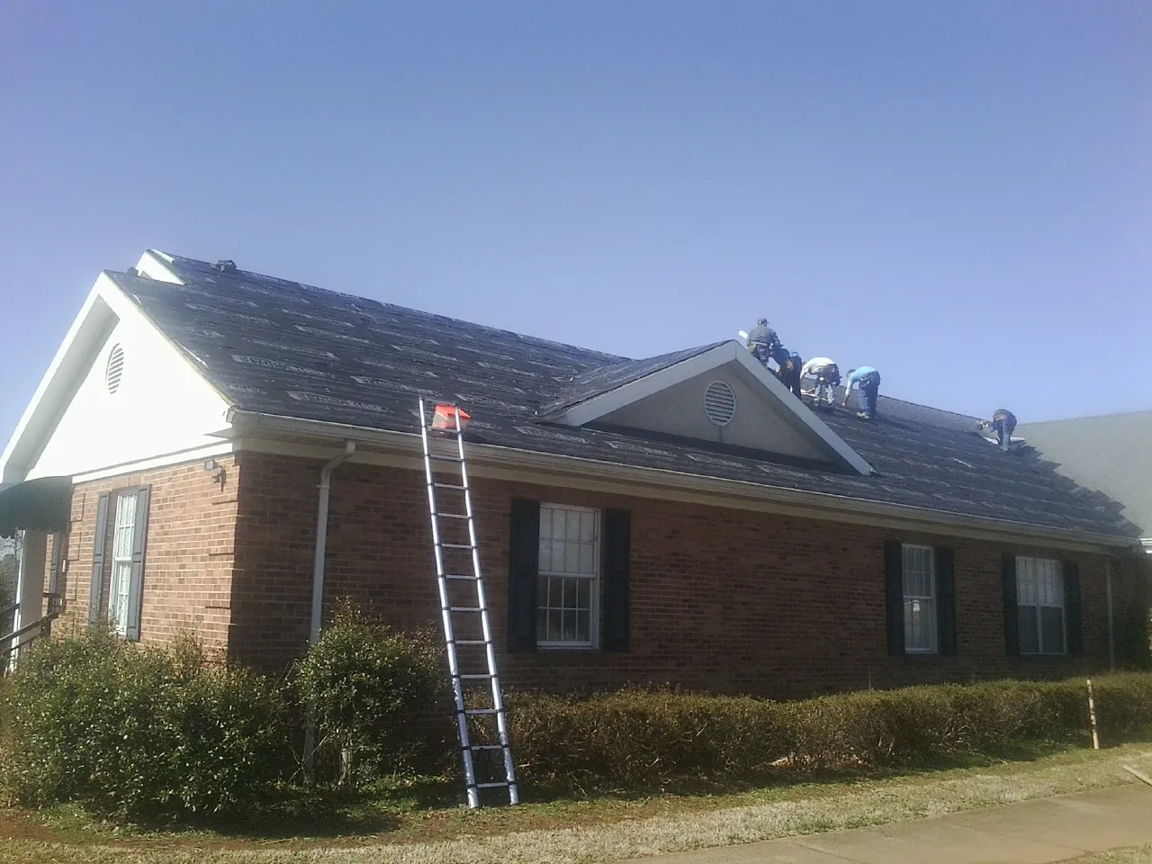 Crew tearing shingles off a brick ranch roof with a ladder set — Emergency Roof Repair work by our crew in Gerlaw