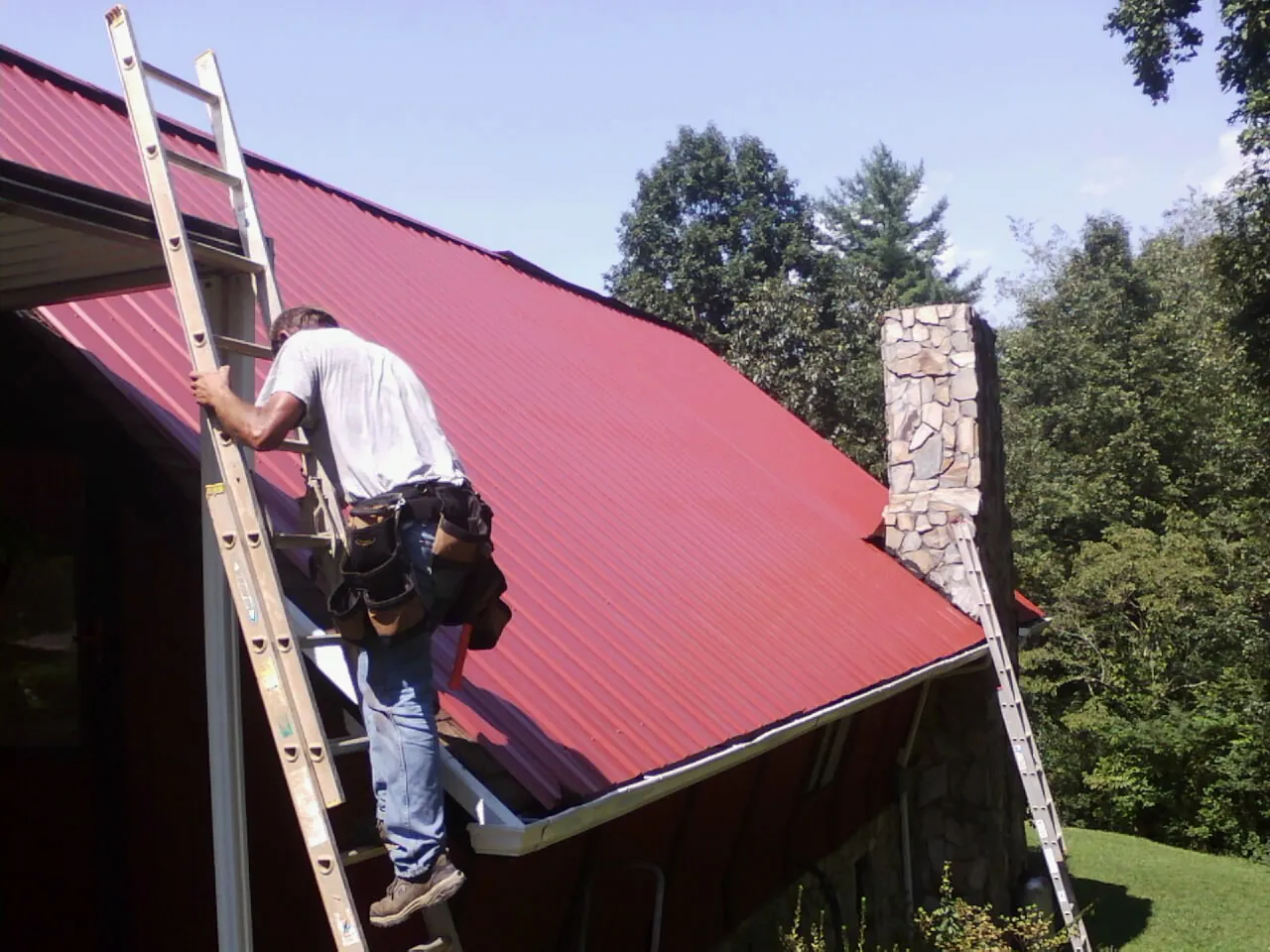 Roofer on a ladder beside a red metal roof and stone chimney — Soffit & Fascia Repair by Roofing Brothers in Gerlaw